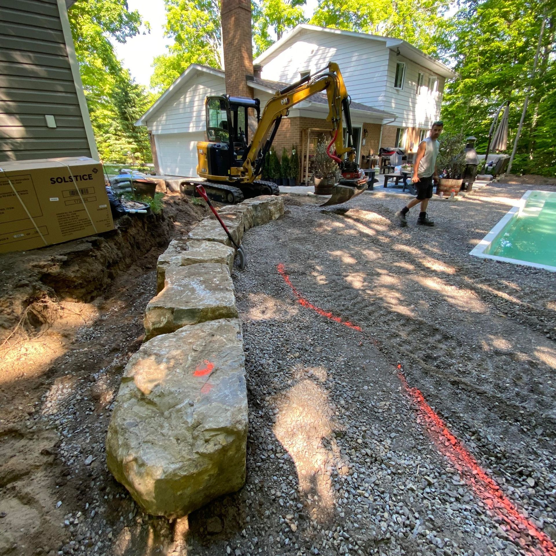 Chantier de construction avec un mur de soutènement en pierre, une excavatrice et une personne marchant près d'une maison et d'une piscine.