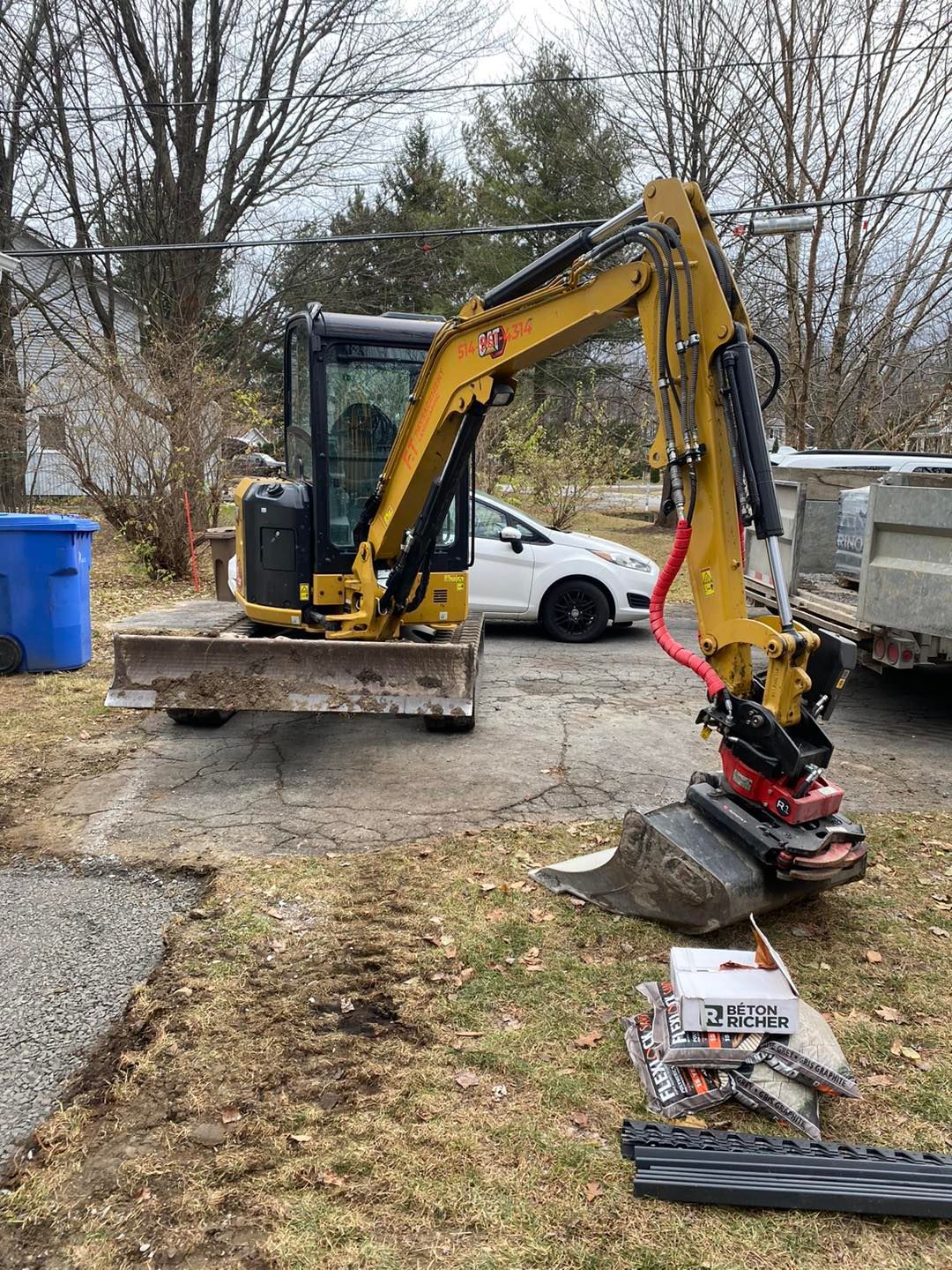 Une pelleteuse jaune est garée sur du gravier près d'une maison, avec ses accessoires et son godet.