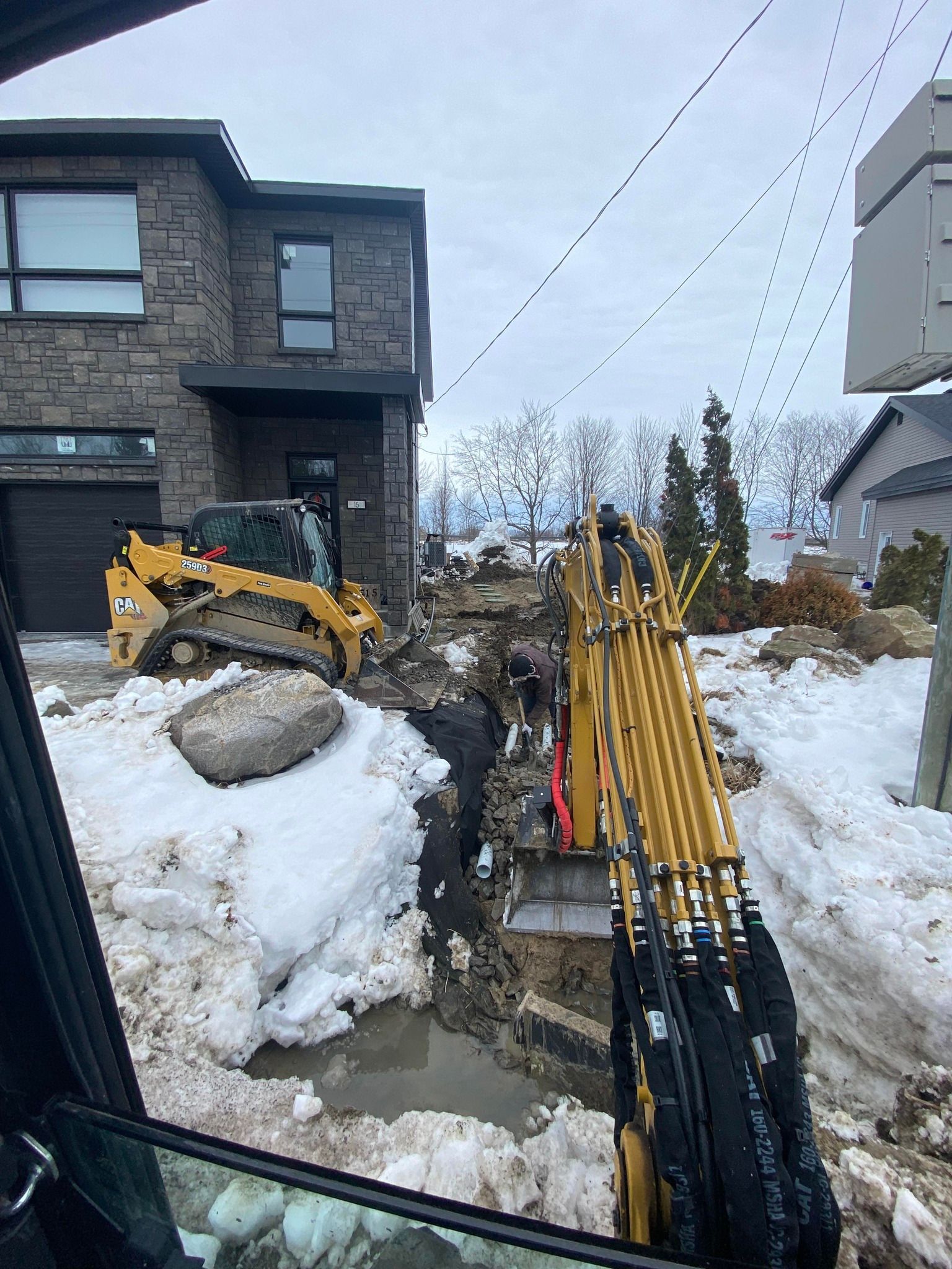 Une excavatrice creuse une tranchée dans la cour avant d'une maison, alors que la neige et des engins de chantier sont présents sur le site.