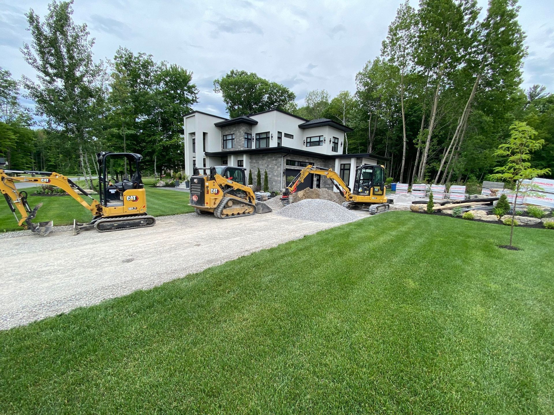 Trois excavatrices sur une allée de gravier devant une maison moderne, travaux d'aménagement paysager en cours.