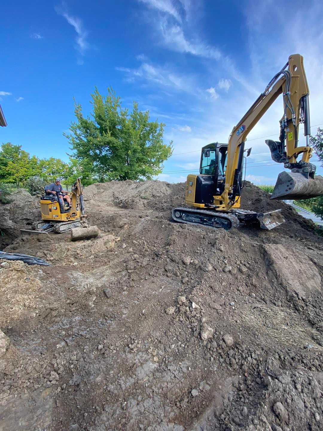 Deux excavatrices jaunes sur une colline de terre sous un ciel bleu, l'une en train de creuser, l'autre au loin.