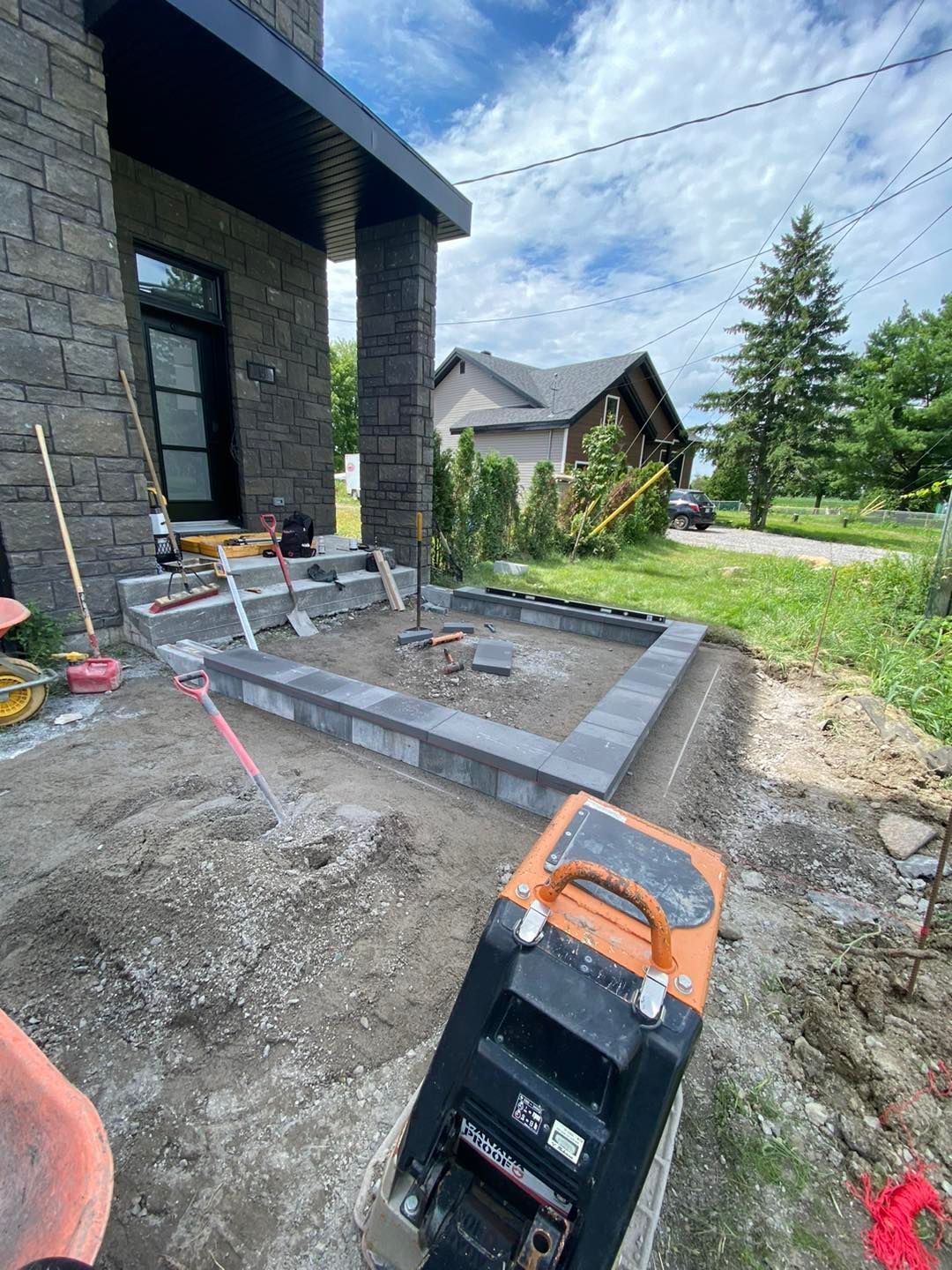Un chantier avec l'entrée d'une maison en pierre grise. Une bordure pavée est visible. Une plaque vibrante est en place.