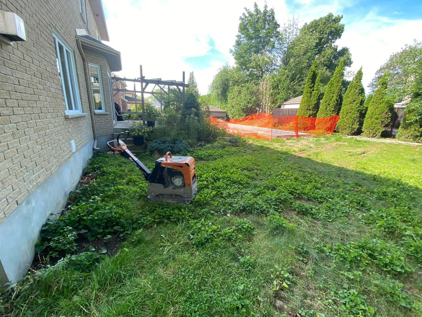 Un jardin avec un compacteur à déchets, des herbes hautes et une maison en briques. Une clôture orange se profile au loin.