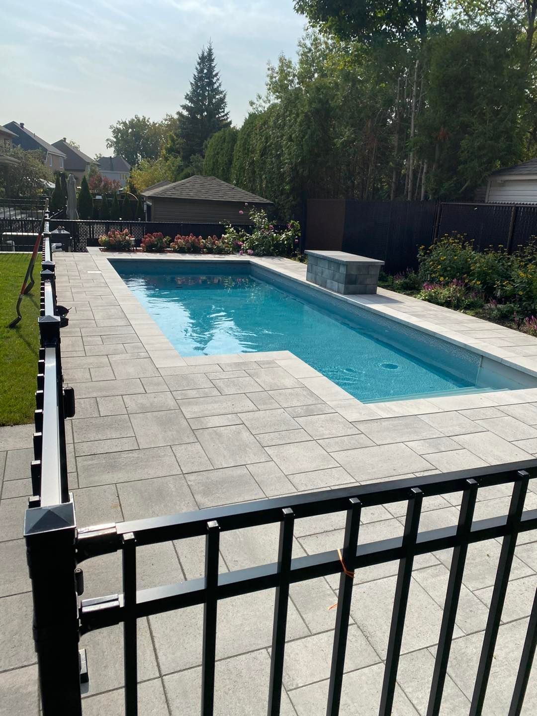 Piscine extérieure à l'eau bleue avec terrasse pavée, entourée d'une clôture noire et d'aménagements paysagers.