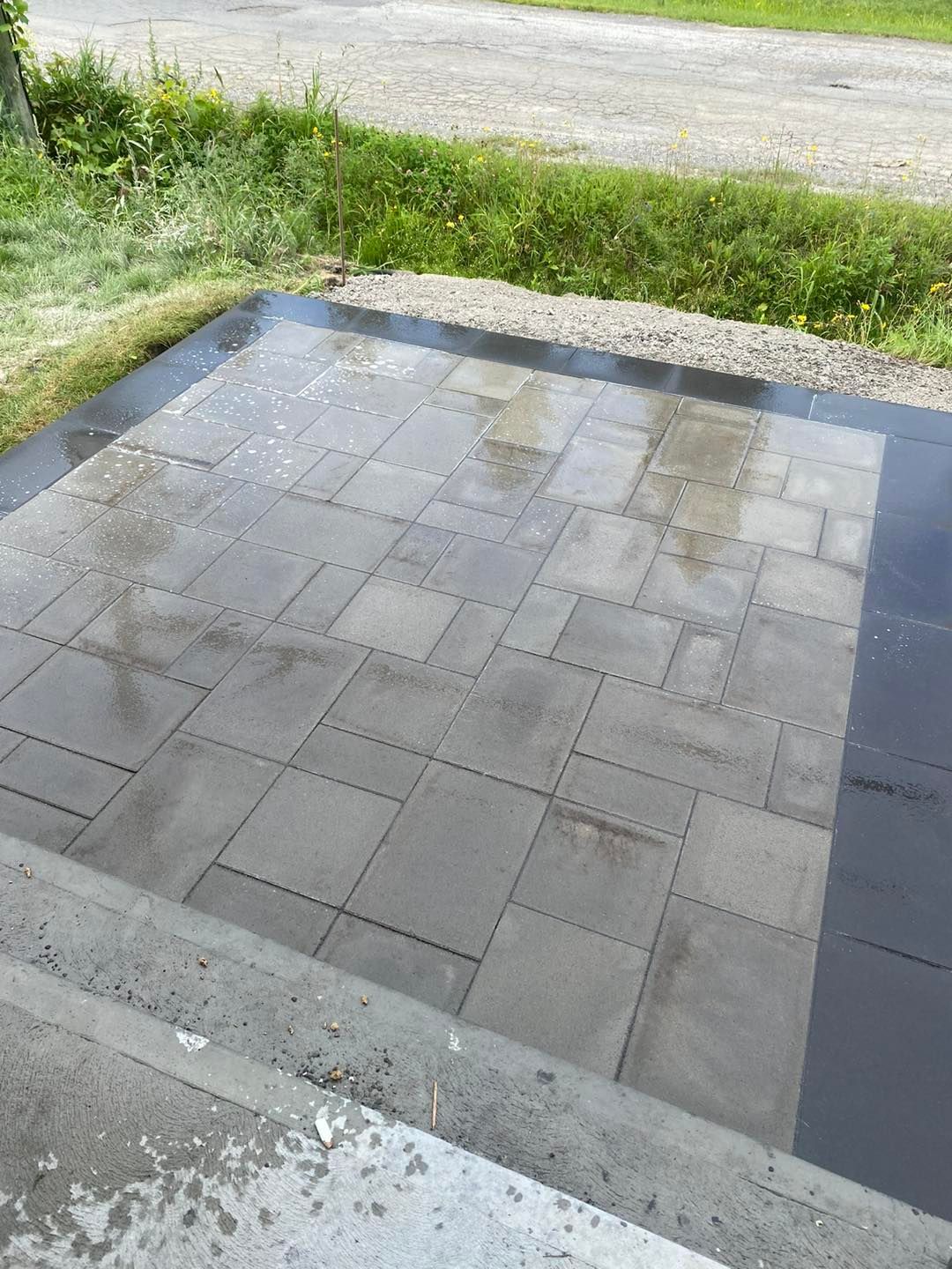 Terrasse en pierre grise et humide avec une bordure plus foncée ; herbe et gravier en arrière-plan.