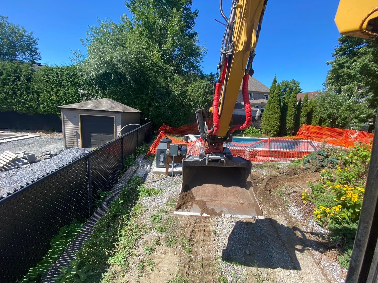 Une pelleteuse creuse près d'une clôture noire, d'un abri de jardin et d'une piscine en construction. Belle journée ensoleillée.