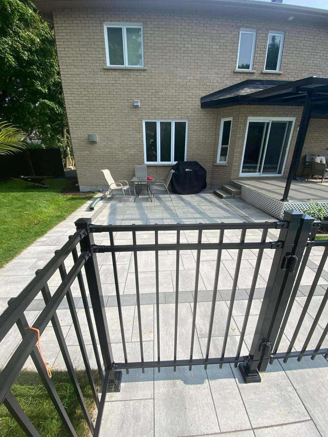 Terrasse arrière avec maison en briques, pavés gris, clôture noire et sièges extérieurs.