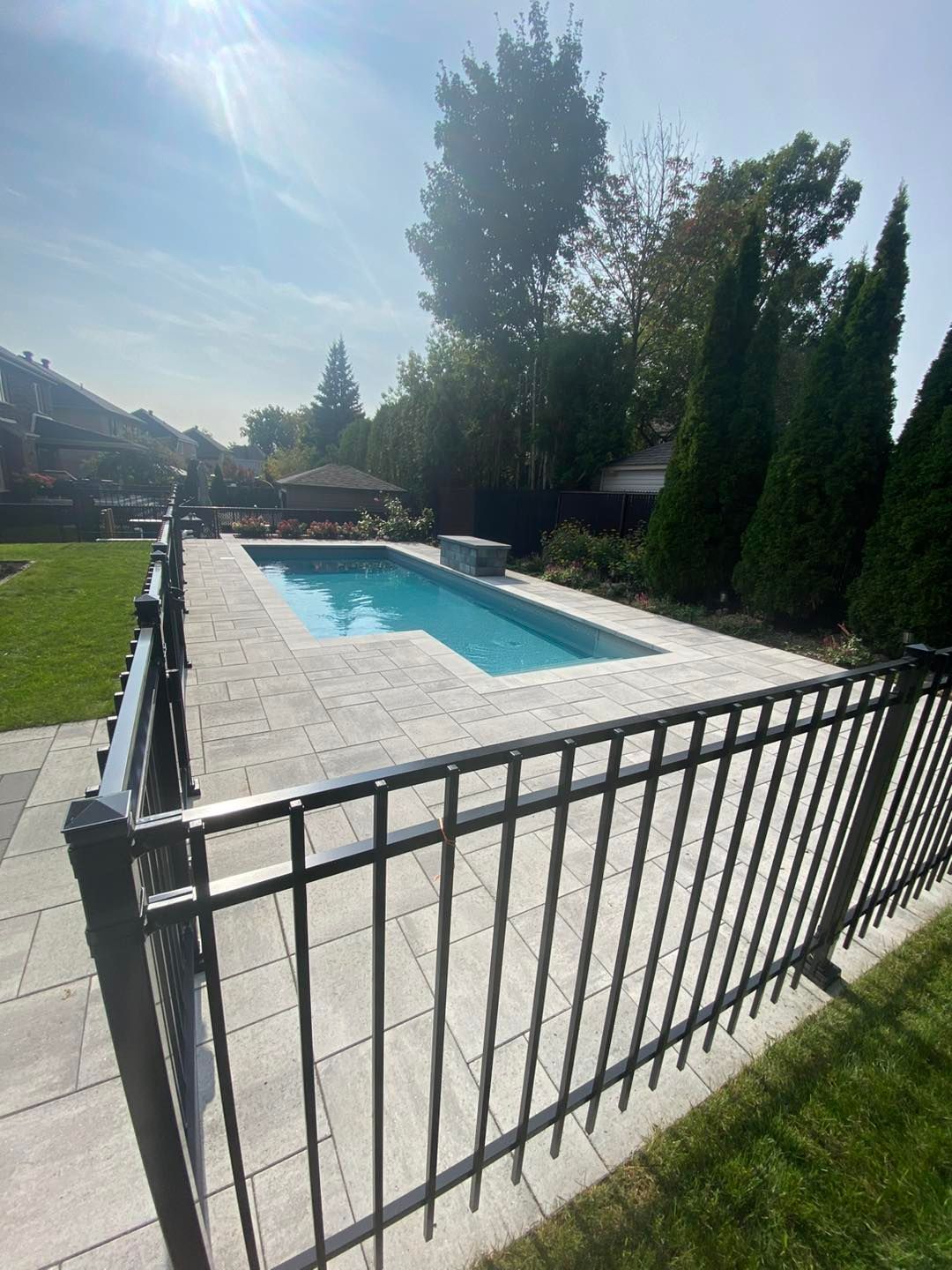 Piscine rectangulaire enterrée entourée de pierres grises. Clôture en métal noir et pelouse verte. Belle journée ensoleillée.