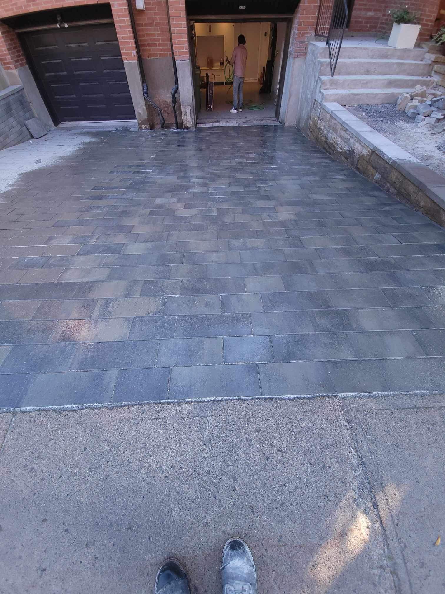 Allée pavée d'un matériau texturé gris foncé menant à l'entrée d'un garage. Une personne se tient à l'intérieur de la porte.