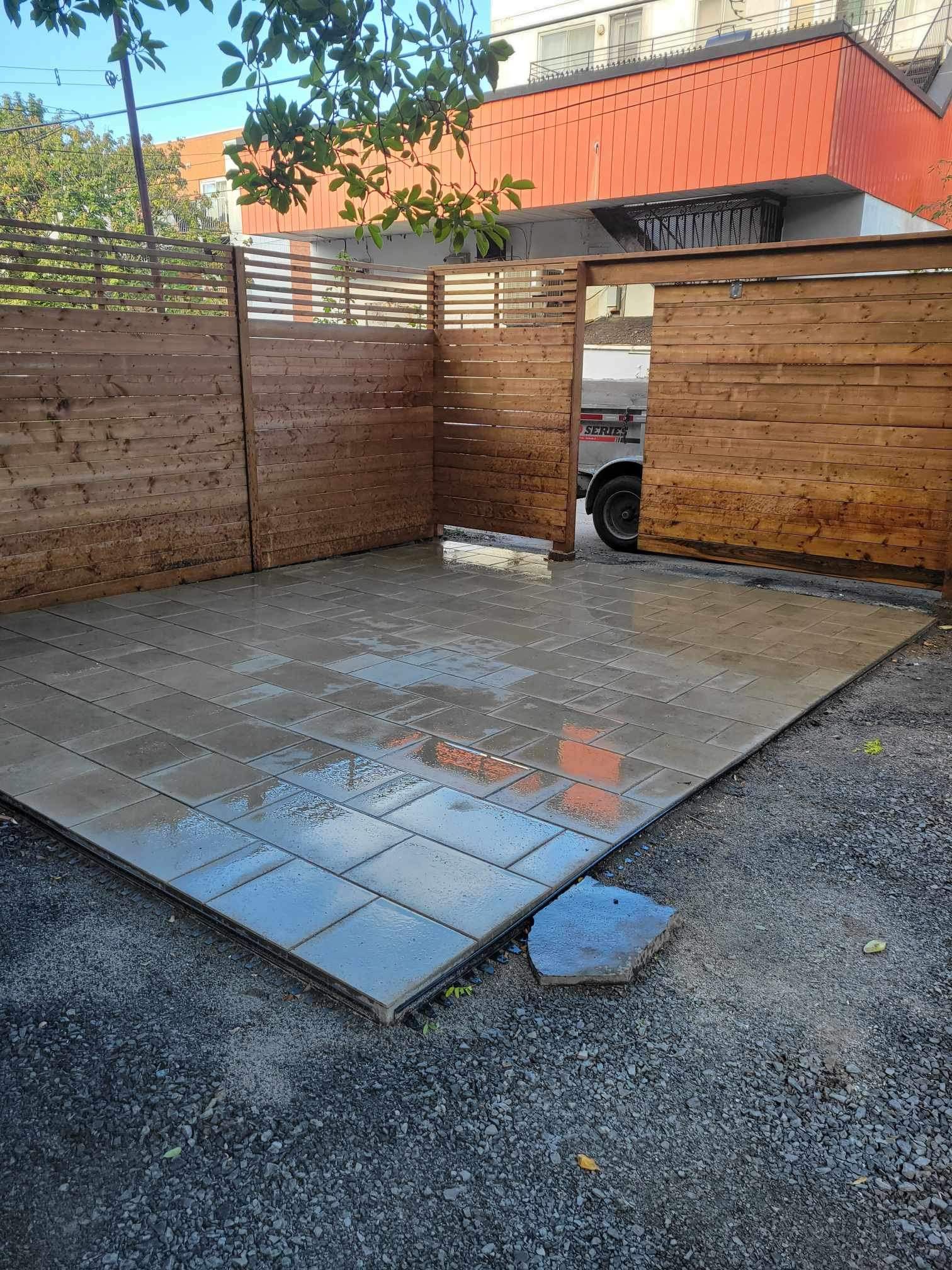 Terrasse extérieure close avec clôture en lattes de bois et dalles grises. Surface humide avec une pierre cassée visible.