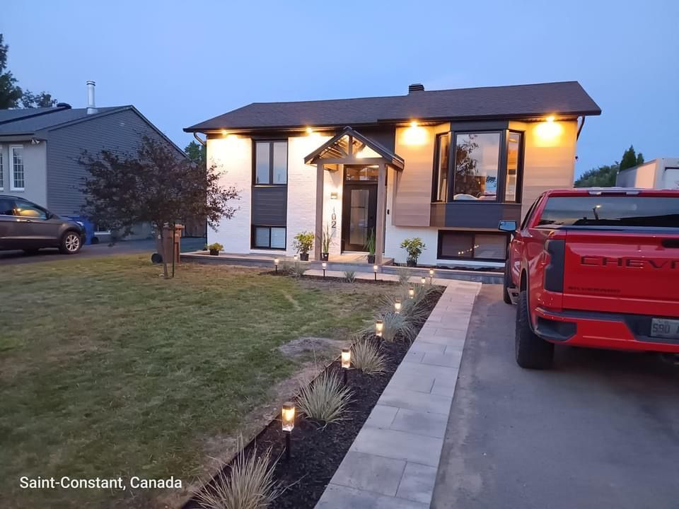 Vue extérieure de la maison au crépuscule, avec un camion rouge dans l'allée. Aménagement paysager et chemin éclairés.