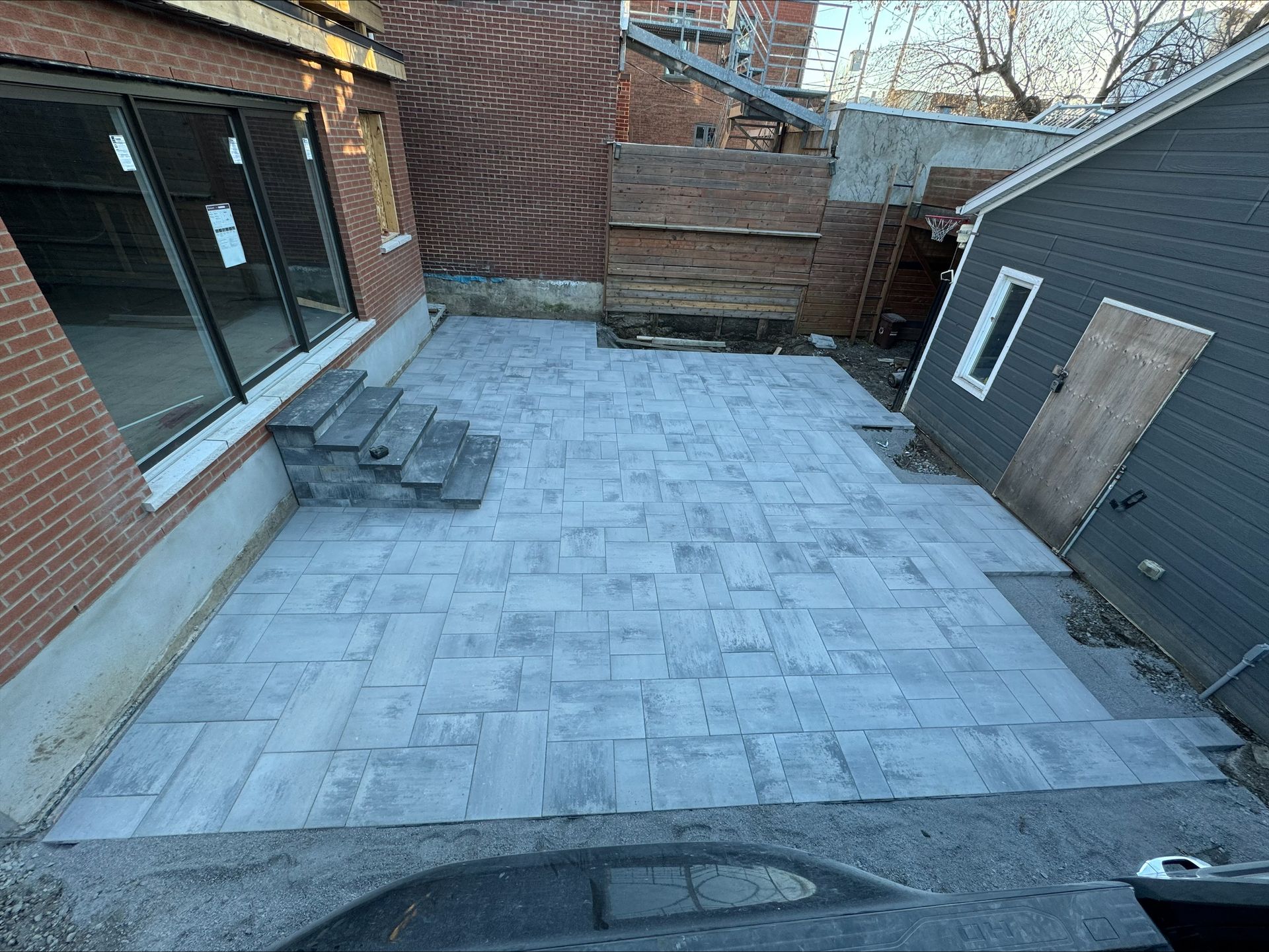 Terrasse en pierre avec marches, jouxtant un bâtiment en briques et un abri de jardin gris.