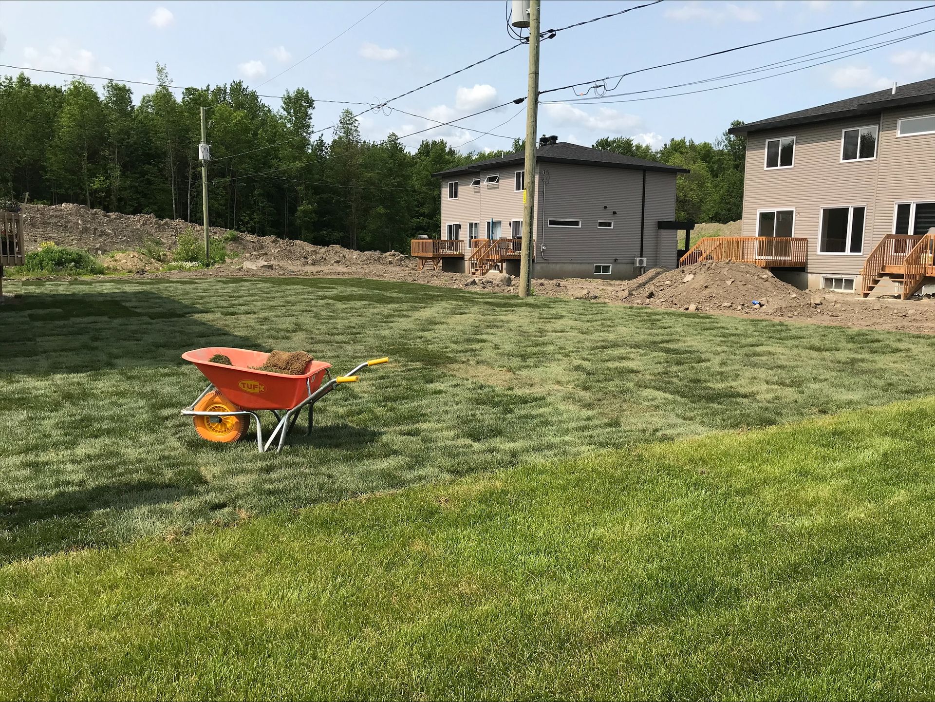 Brouette sur l'herbe, chantier de construction avec maisons à deux étages, tas de terre et arbres.