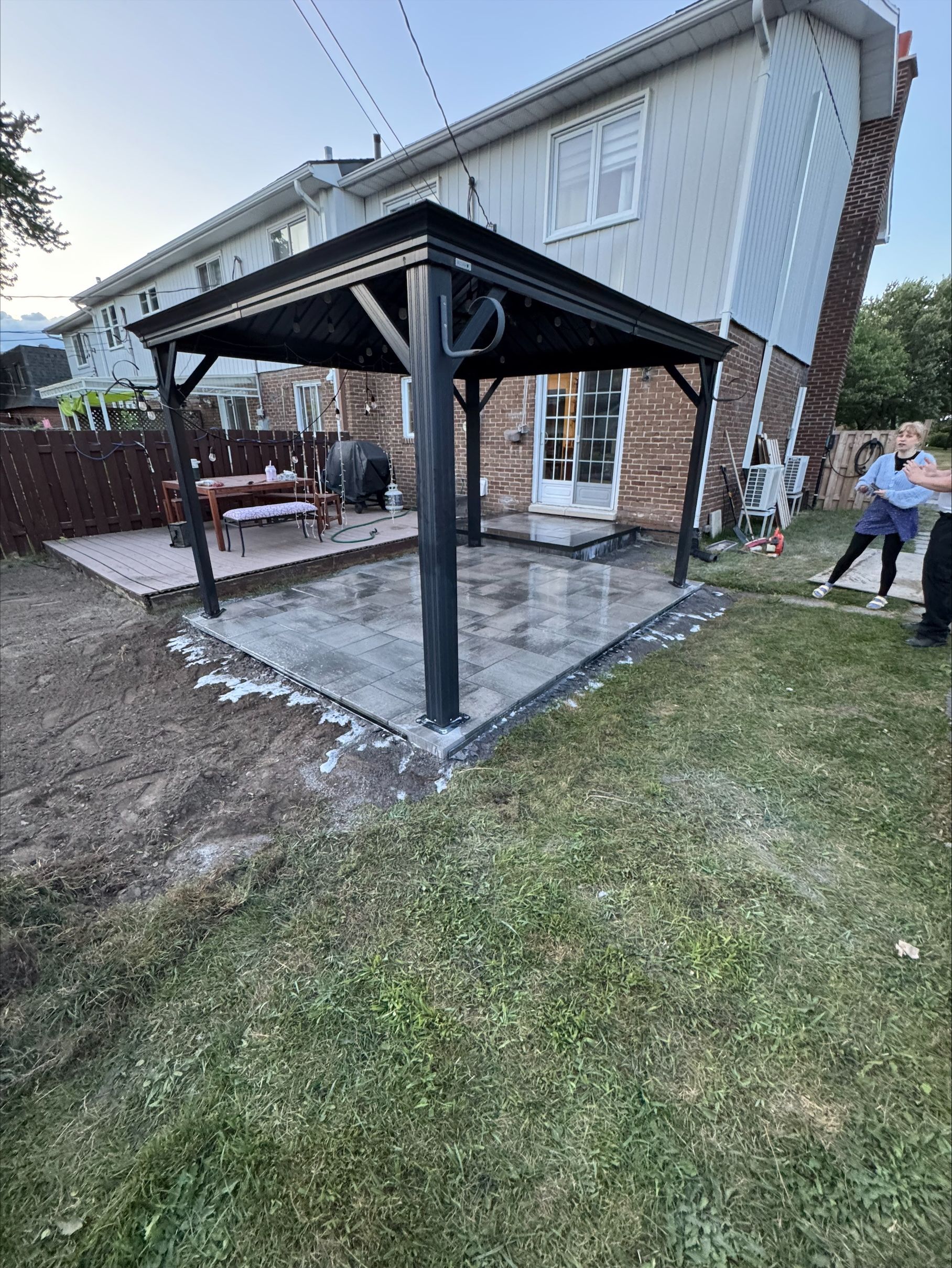 Gazebo noir sur une terrasse en béton dans un jardin ; deux personnes près d’une maison, de l’herbe et d’une clôture.