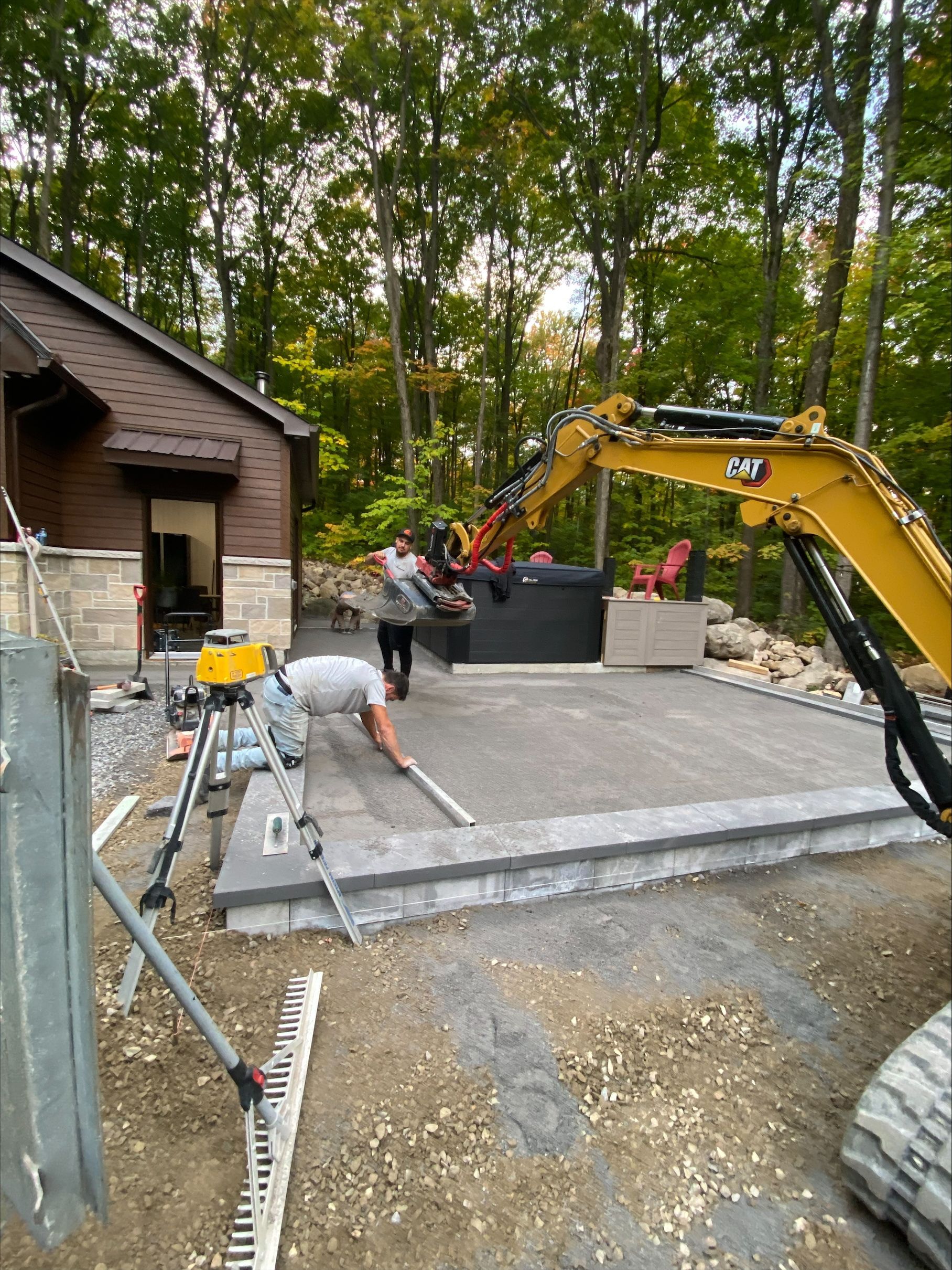Des ouvriers du bâtiment posent des pavés pour une terrasse ; une excavatrice est visible en arrière-plan, des arbres et un bâtiment sont également visibles.