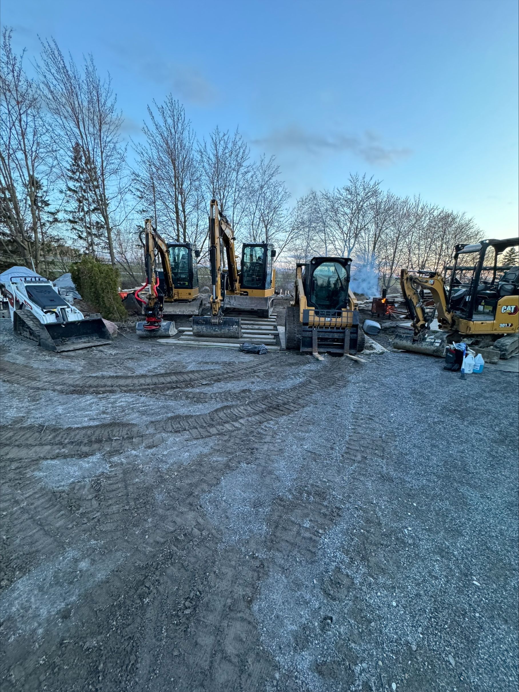 Des engins de chantier, dont des pelles mécaniques et une chargeuse compacte, sont stationnés sur un terrain gravillonné sous un ciel bleu.