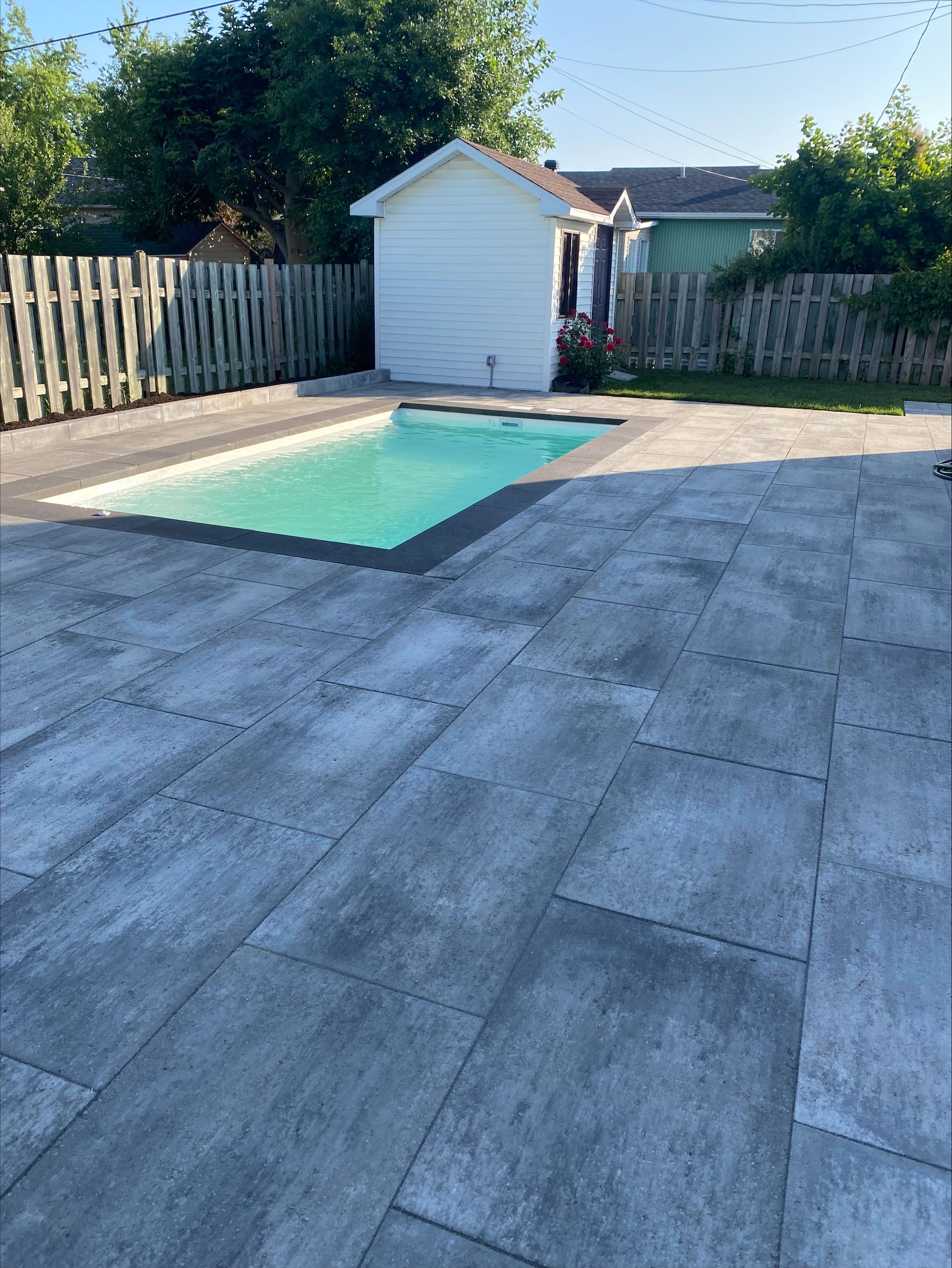 Jardin avec piscine et dalles grises ; petit abri blanc en arrière-plan.