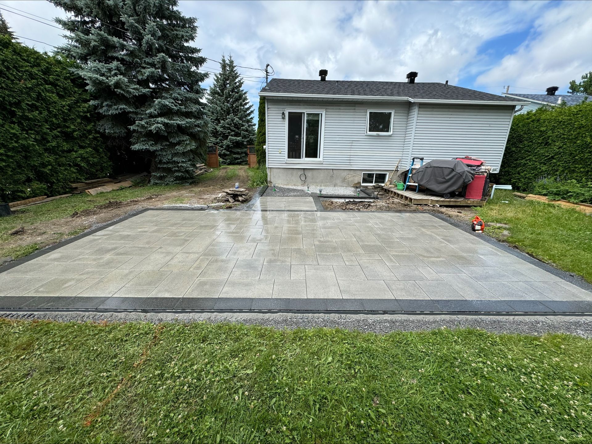 Terrasse arrière avec pavés gris et bordure de blocs plus foncés, jouxtant une maison avec pelouse.