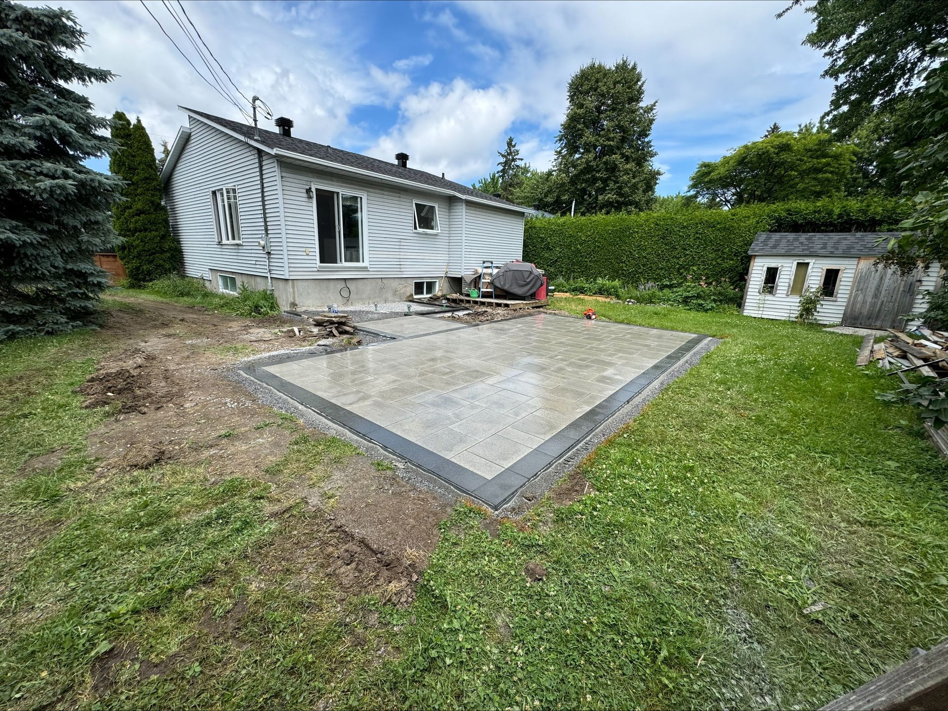 Jardin avec terrasse en béton, maison, abri de jardin et végétation environnante sous un ciel nuageux.