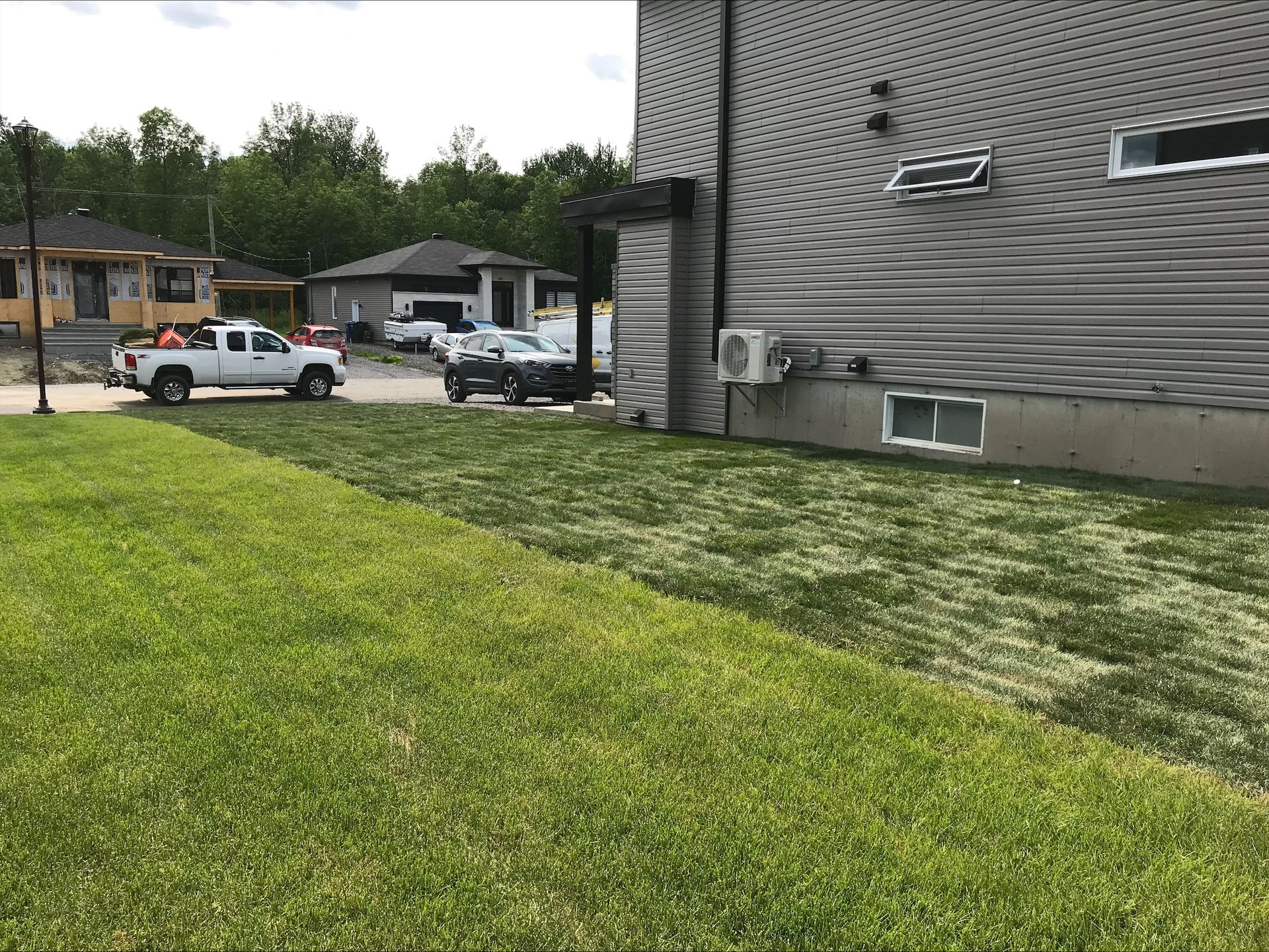 Un camion blanc est garé sur l'herbe à côté d'une maison. D'autres véhicules et bâtiments se dressent à l'arrière-plan, par une journée ensoleillée.