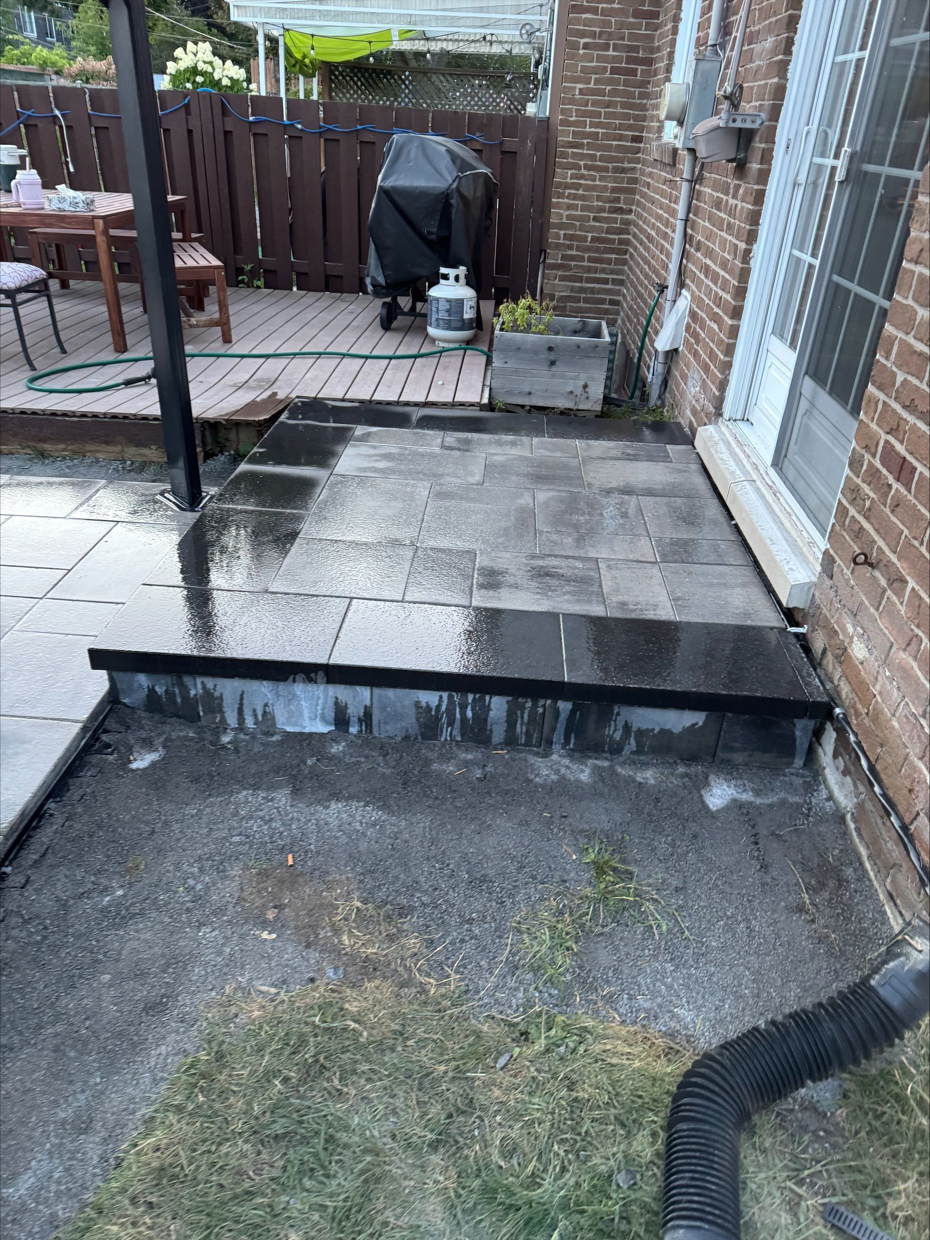 Terrasse en pierre humide avec marches, jouxtant un bâtiment en briques. Gouttière de drainage au premier plan. Clôture marron, barbecue visible.