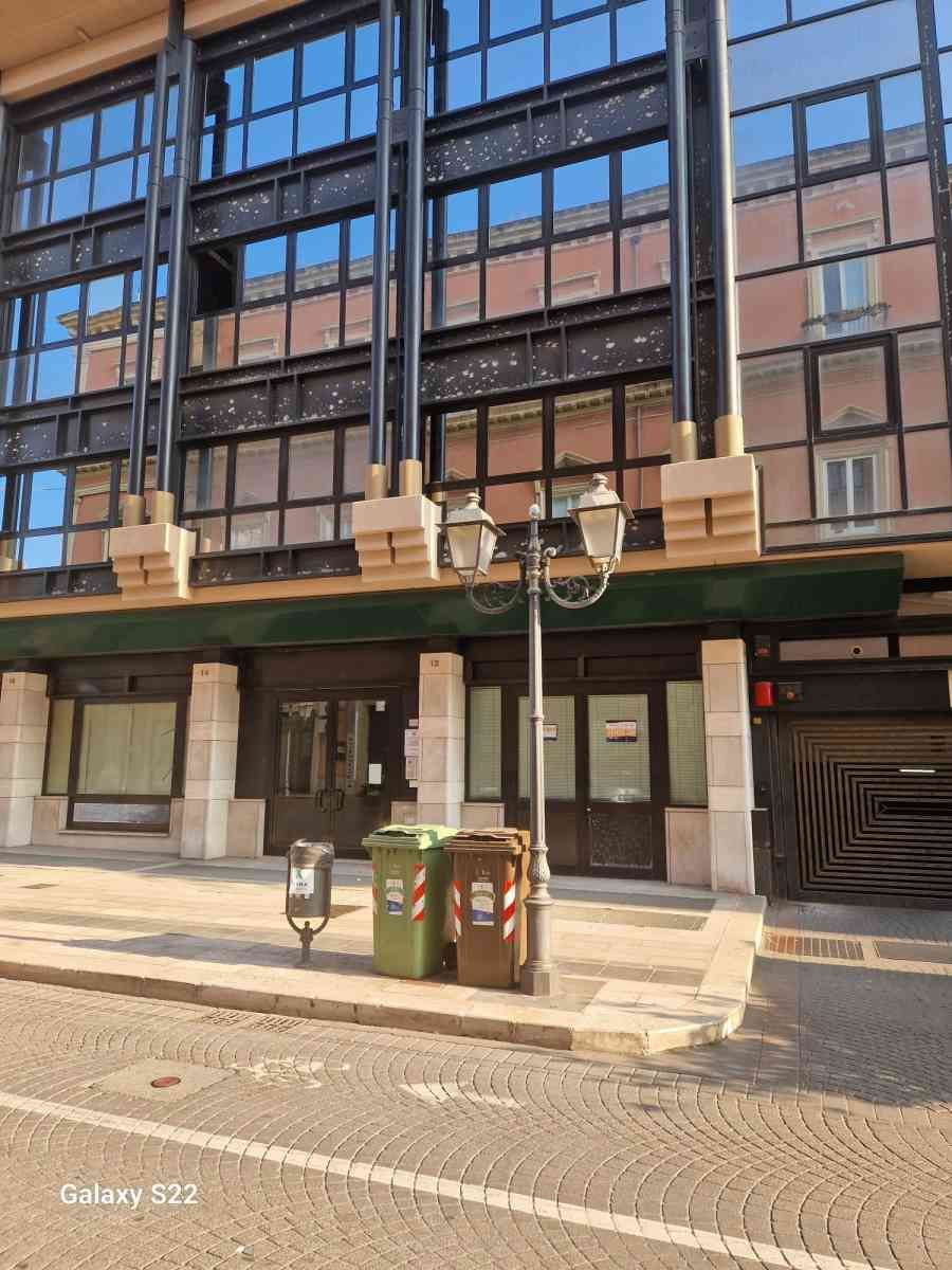 Vista a livello stradale di un moderno edificio con facciata in vetro, tenda da sole verde e vetrine su una strada acciottolata.