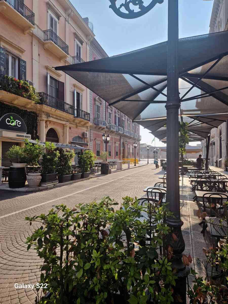 Terrazza soleggiata di un caffè in una tranquilla strada europea con edifici rosa e piante in vaso.