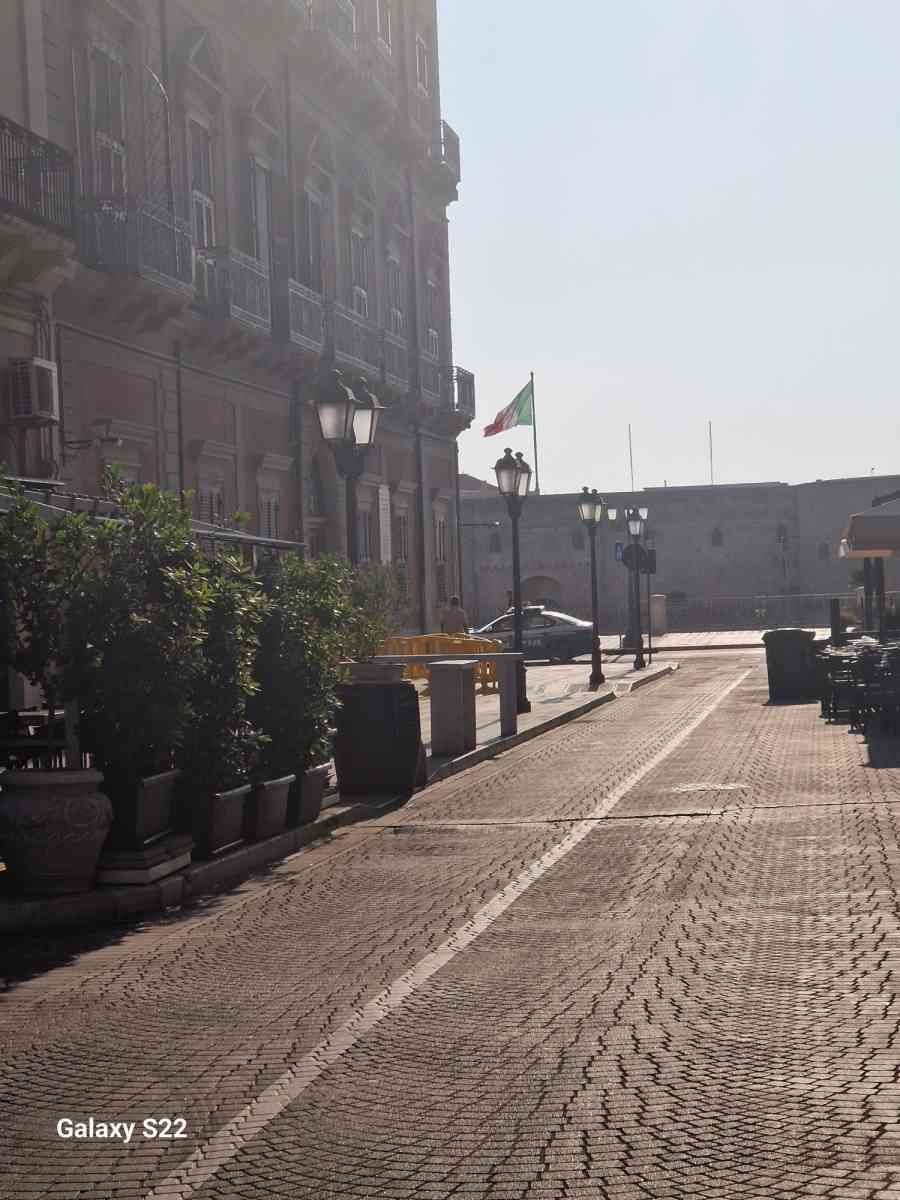 Una strada lastricata di ciottoli illuminata dal sole, accanto a vecchi edifici, piante in vaso e una bandiera su una tranquilla passeggiata sul lungomare.