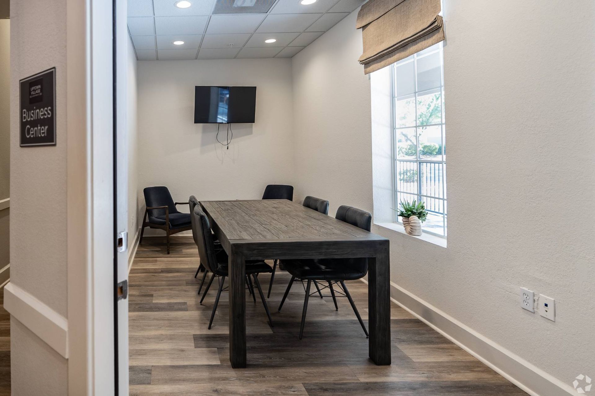 Community business center with a large wooden conference table, black chairs, wall-mounted TV, and a window at Uptown Village Apartments in Gainesville, FL.