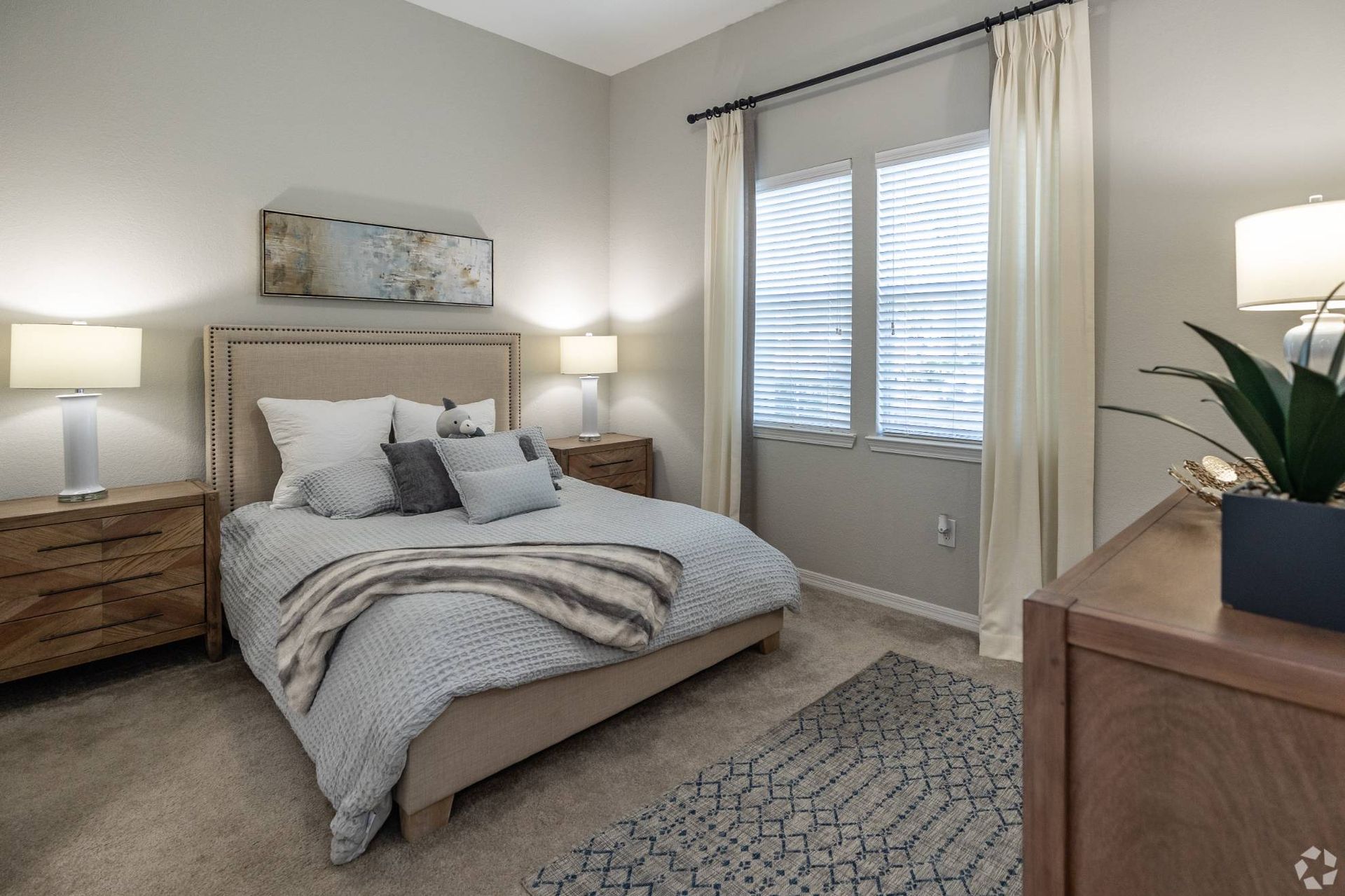 Bedroom in an apartment with a beige upholstered bed, two nightstands, lamps, and window with blinds at Uptown Village Apartments in Gainesville, FL.