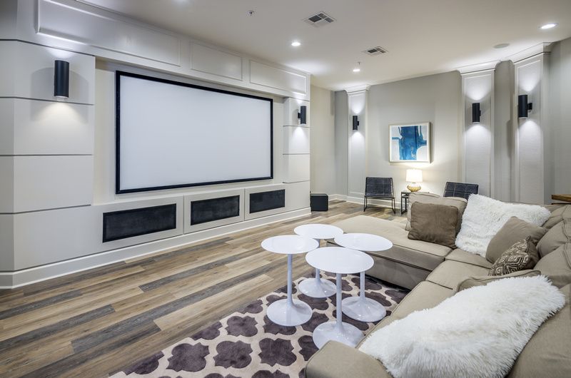 Modern community lounge with a large wall-mounted screen, beige sectional sofa, and white round tables at Uptown Village Apartments in Gainesville, FL.