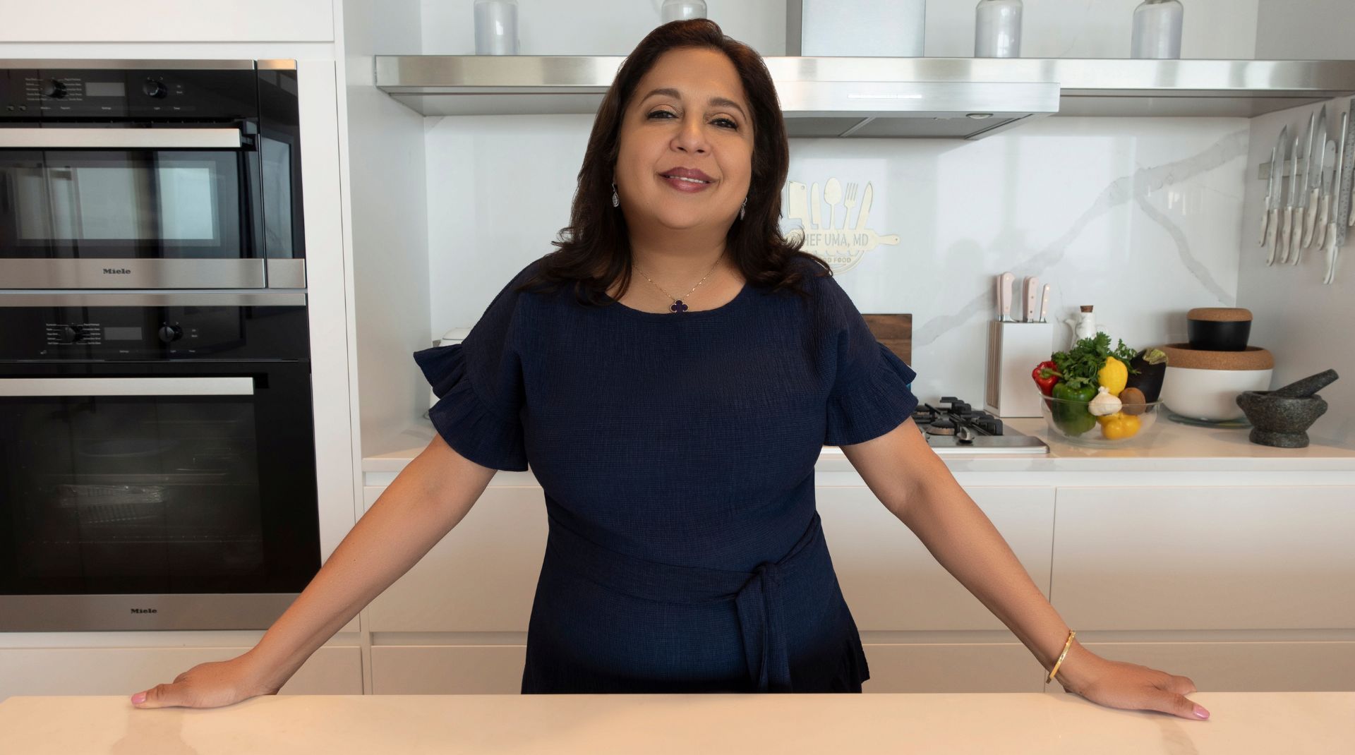 Dr. Uma Naidoo standing in a kitchen, smiling confidently with fresh ingredients in the background.
