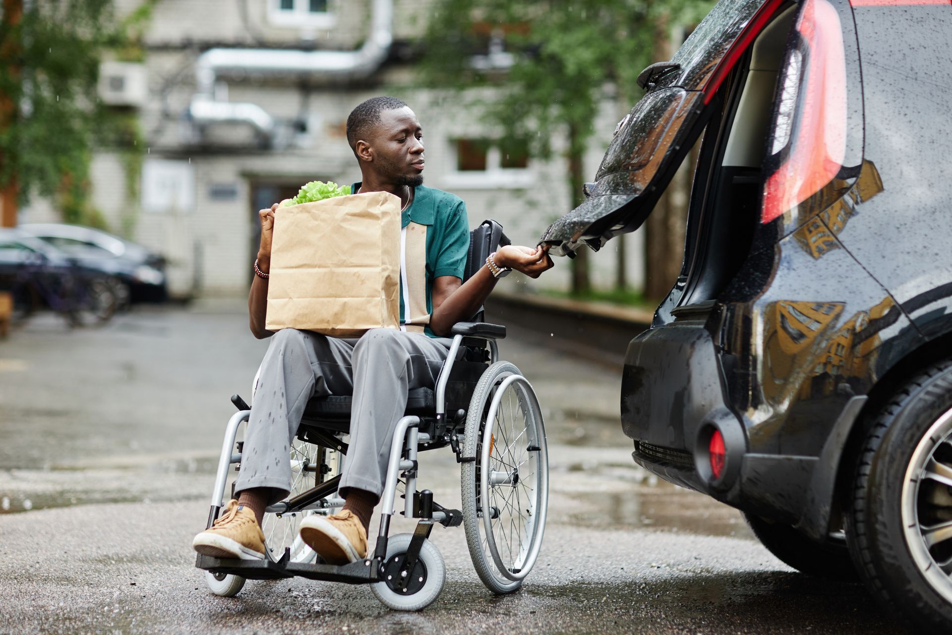 Person in a wheelchair reaches toward the open trunk of a black car on a wet street. Person in a wheelchair reaches toward the open trunk of a black car on a wet street.