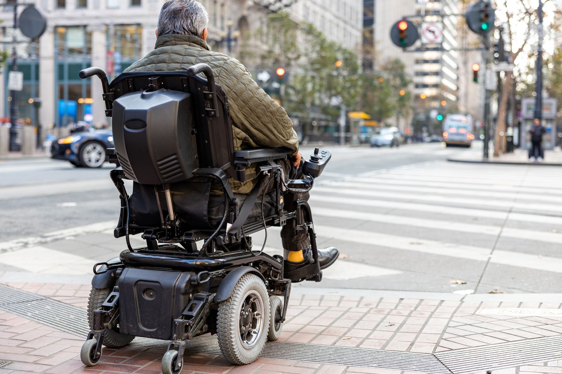 Person in a motorized wheelchair at a city crosswalk, viewed from behind. Person in a motorized wheelchair at a city crosswalk, viewed from behind.