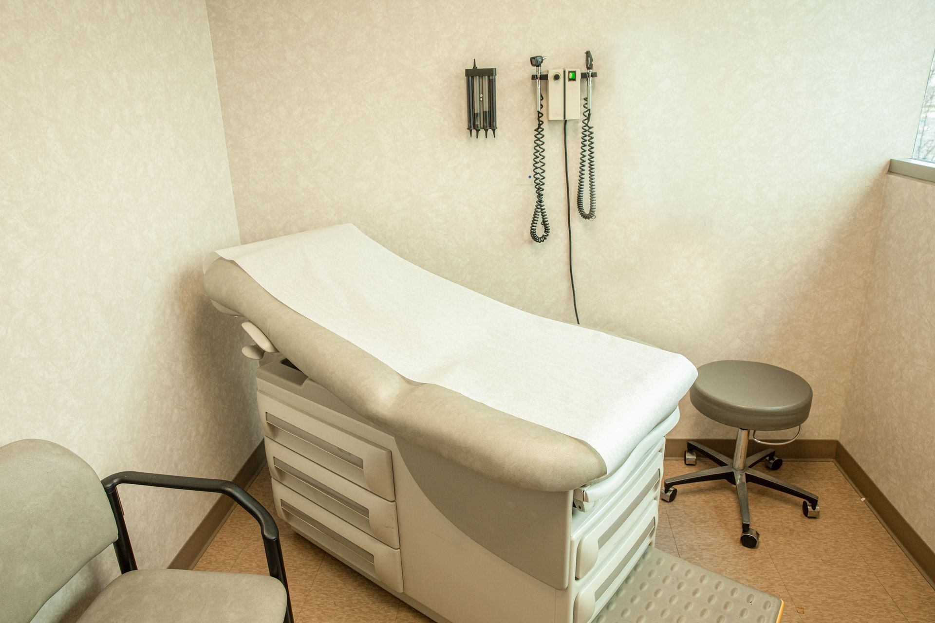 Medical exam room with padded exam table, stool, chair, and wall-mounted tools