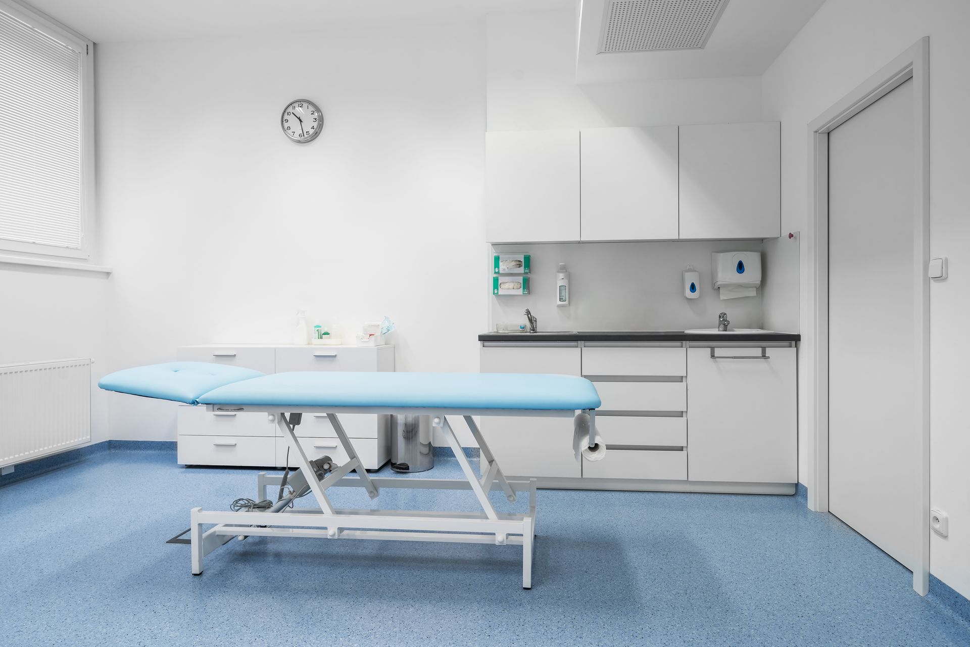 Empty medical exam room with examination table, sink, cabinets, and wall clock