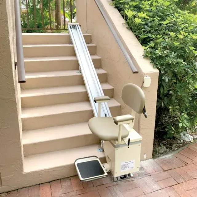 Outdoor stairlift with beige seat and rail mounted along a brick staircase beside greenery.