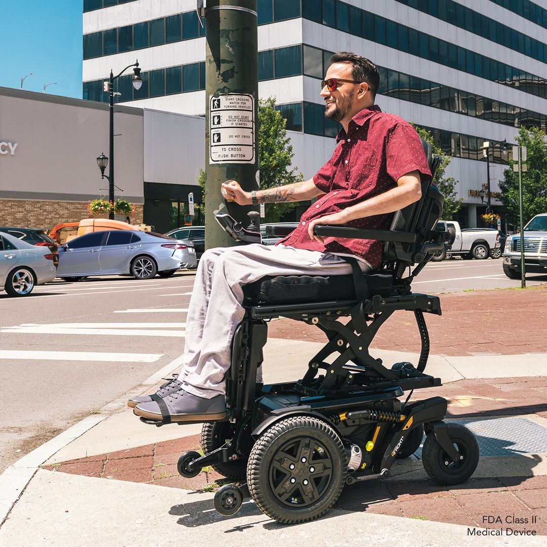 Person riding a black-and-yellow wheelchair on a sunlit beach path, wearing a white shirt and pants