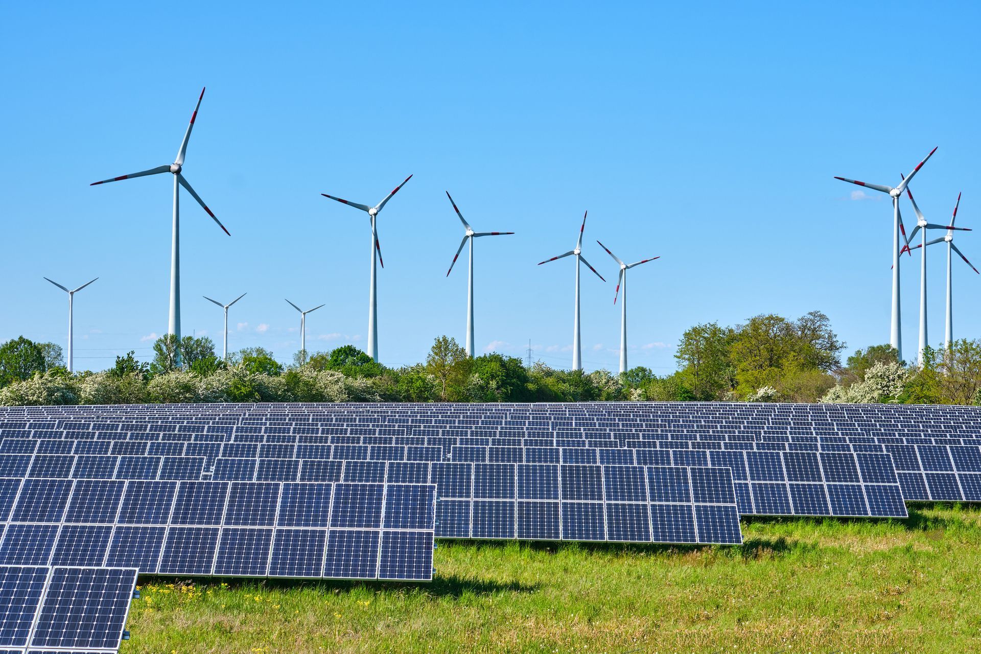 Solar panels and wind turbines in a grassy field under a blue sky, representing renewable energy sources. Solar panels and wind turbines in a grassy field under a blue sky, representing renewable energy sources.