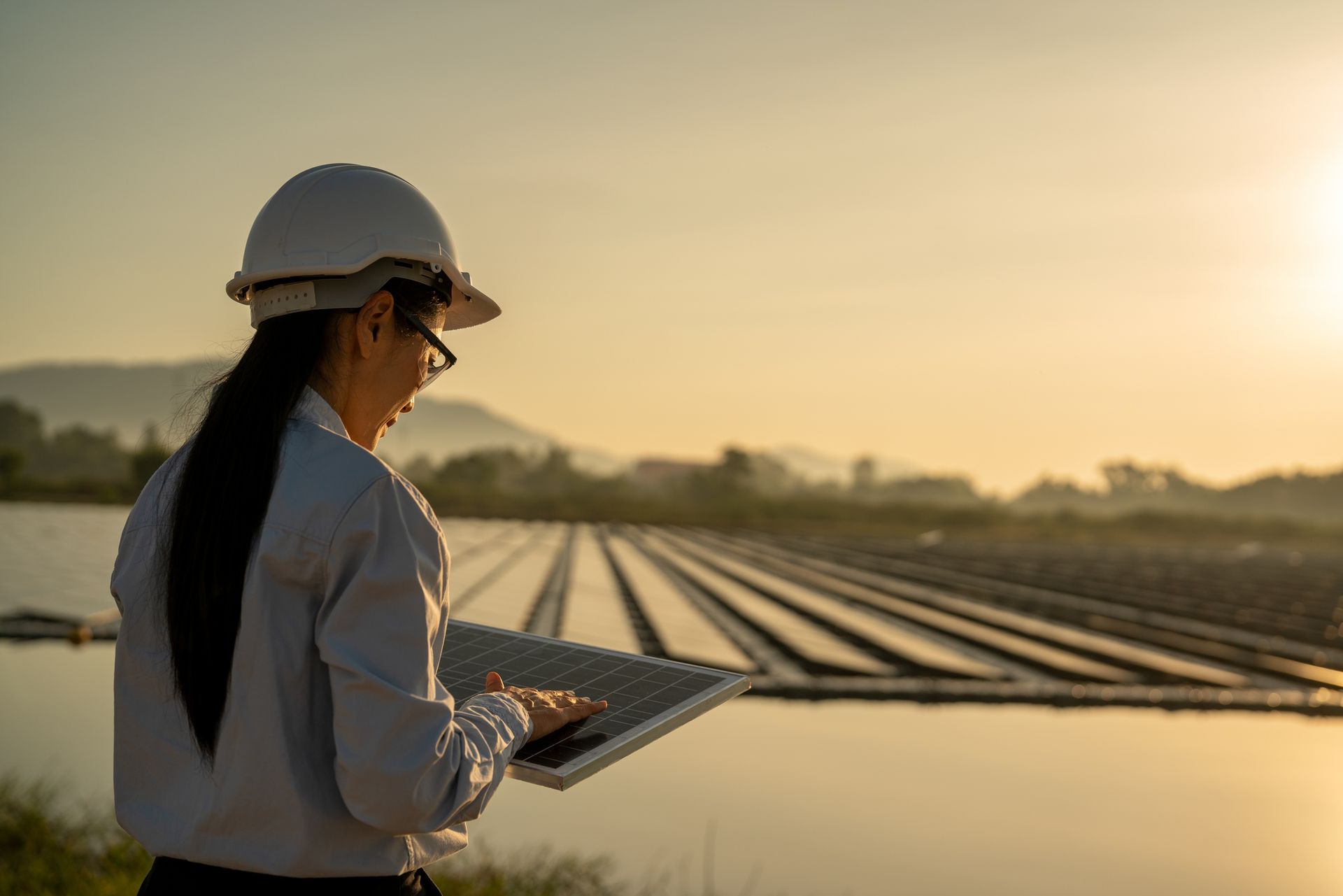 Woman with hard hat and glasses uses a tablet in front of a solar panel field at sunset. Woman with hard hat and glasses uses a tablet in front of a solar panel field at sunset.