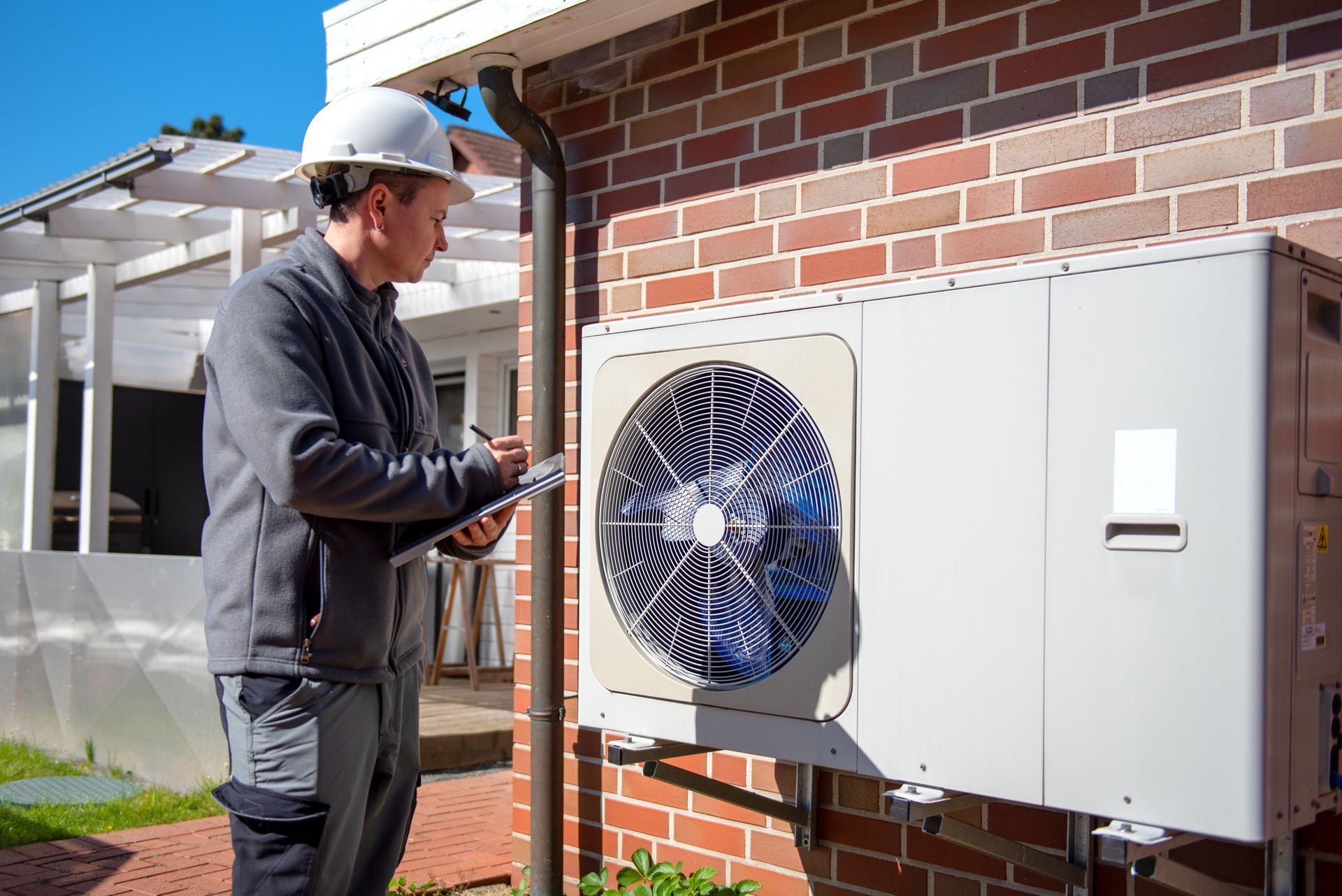 HVAC technician in hard hat inspecting a white air conditioning unit mounted on a brick wall, taking notes. HVAC technician in hard hat inspecting a white air conditioning unit mounted on a brick wall, taking notes.