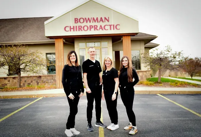 Four staff members pose outside Bowman Chiropractic office with red signage and parking lot in front.