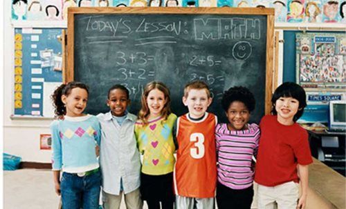 Five children standing in front of a chalkboard in a classroom, smiling and holding hands