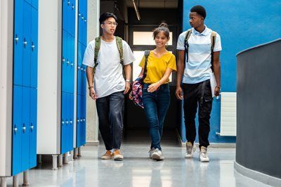 Three students walking down a school hallway with blue lockers, carrying backpacks and books.
