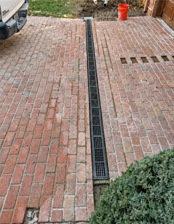 Brick driveway with a long narrow drainage grate down the center and a red bucket near the top