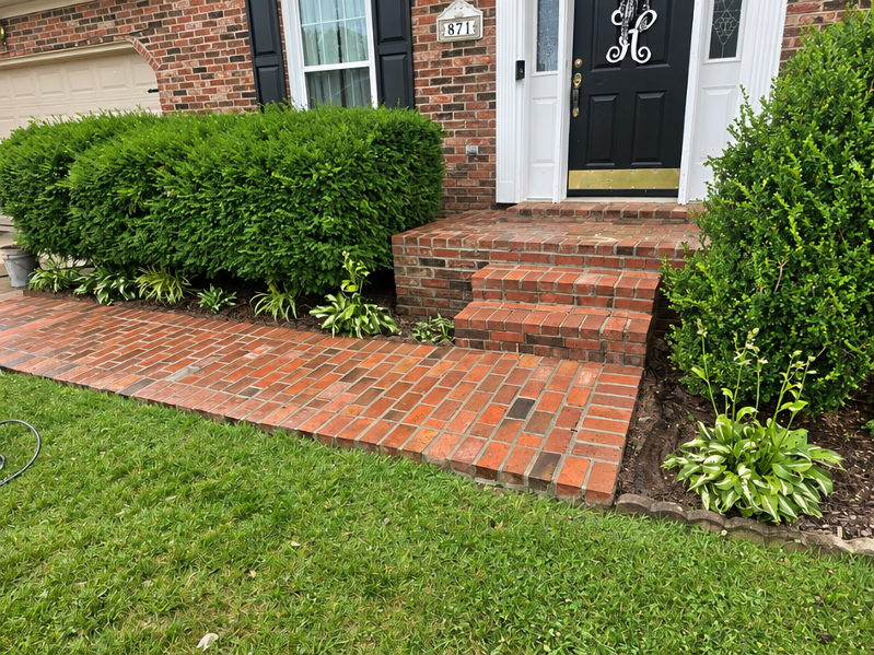 Brick front steps and walkway leading to a white door, bordered by green shrubs and lawn.