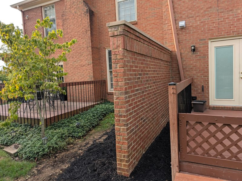 Brick privacy wall beside a townhouse courtyard with black railing, green shrubs, and a wood lattice fence