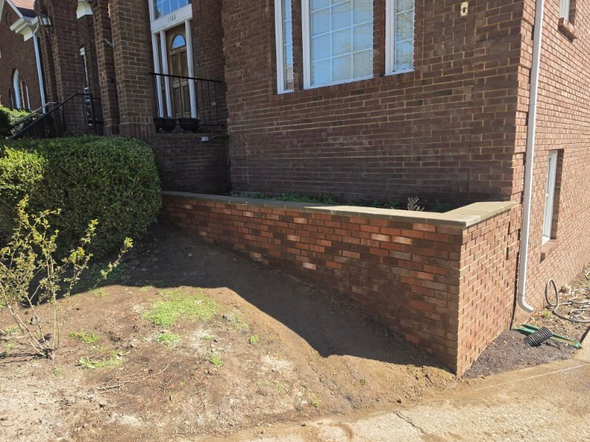 Brick house entrance with a raised front wall, steps, and dirt yard under bright sunlight