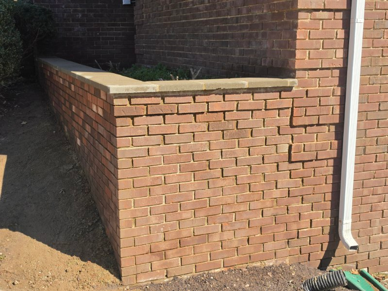 Brick planter wall beside a house, with a white downspout and a garden hose nearby.