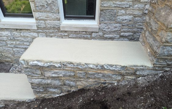 Stone house foundation with concrete ledges and two windows above a sloped dirt edge